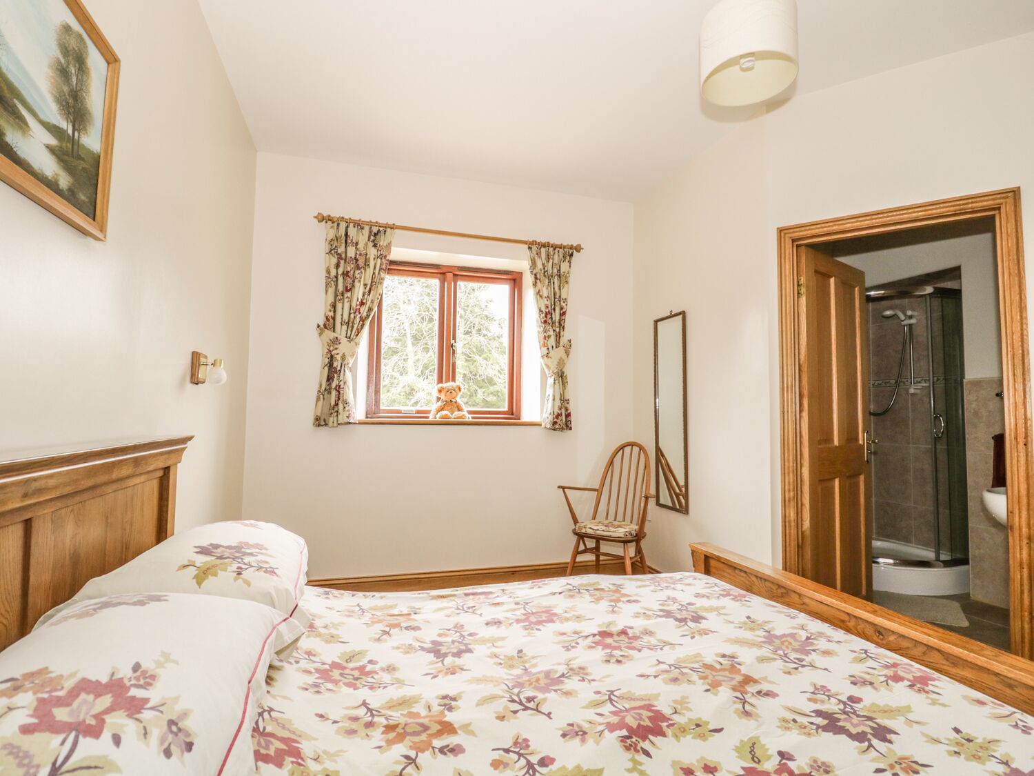 Ein Schlafzimmer mit Bett, Sessel, Fenster mit Vorhängen, Spiegel und Duschbereich.