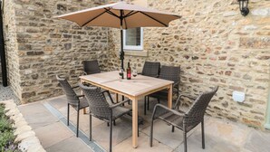 A stone wall patio with a wooden table and wicker chairs, a beige umbrella, and a bottle of red wine. - Cross Cottage (Durham)