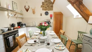 A dining area with a table set for four, surrounded by wooden chairs. A kitchen is visible to the left with modern appliances. - Cross Cottage (Durham)
