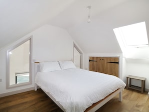 Ein Schlafzimmer mit schrägen Wänden, einem Bett mit weißer Bettwäsche, Holzboden, einem kleinen Nachttisch und einem Dachfenster.