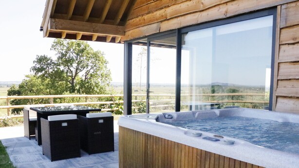 Eine Holzterrasse mit Whirlpool, Sitzgelegenheiten im Freien und einem großen Fenster mit Blick auf eine reizvolle Landschaft.