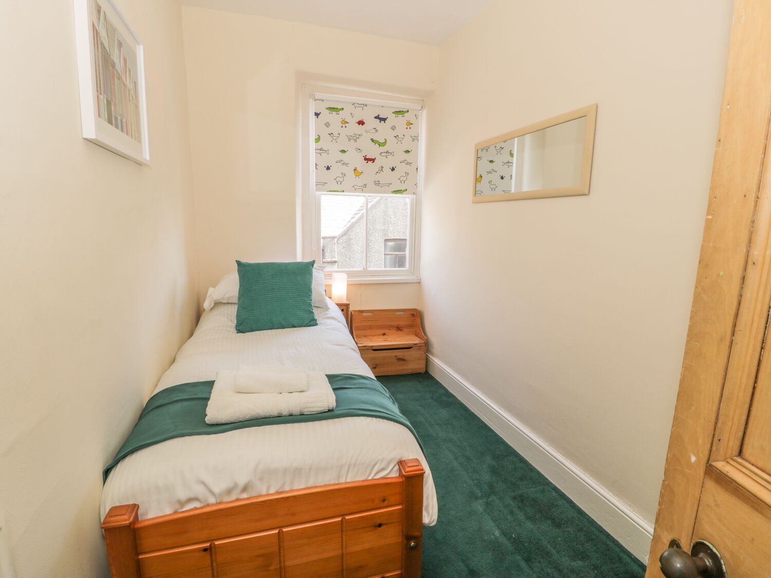 Une petite chambre à coucher accueillante, avec un lit simple, une fenêtre ornée d’un rideau à motifs, une table de chevet en bois et un miroir accroché au mur.