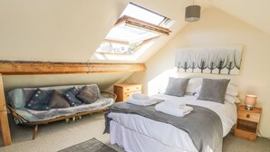 Un dormitorio con tragaluces, una cama con ropa blanca, un sofá gris con almohadones de colores y una mesita de noche de madera con una lámpara.