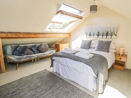 Un dormitorio con tragaluces, una cama con ropa blanca, un sofá gris con almohadones de colores y una mesita de noche de madera con una lámpara.