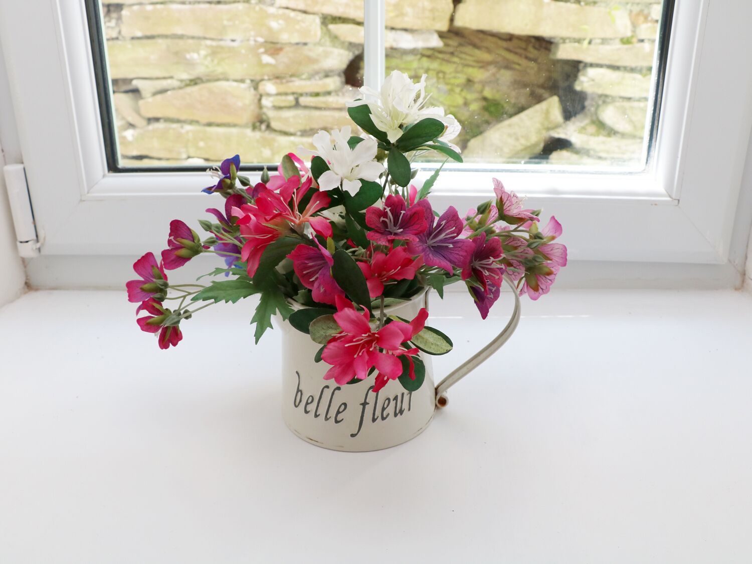 Un vase en céramique blanche, orné de l’inscription « belle fleur », est posé sur un rebord de fenêtre.