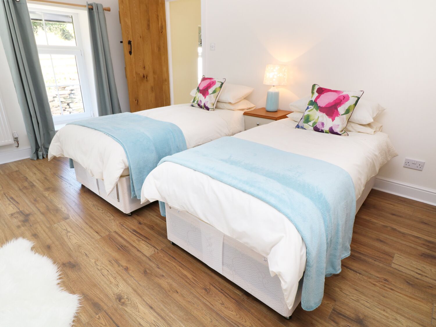 A bedroom with two beds, wooden flooring, a nightstand with a lamp, and a window with curtains.