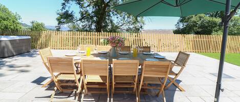Een terras buiten met een ronde eettafel, omringd door houten stoelen, een groene parasol en een jacuzzi op de achtergrond.