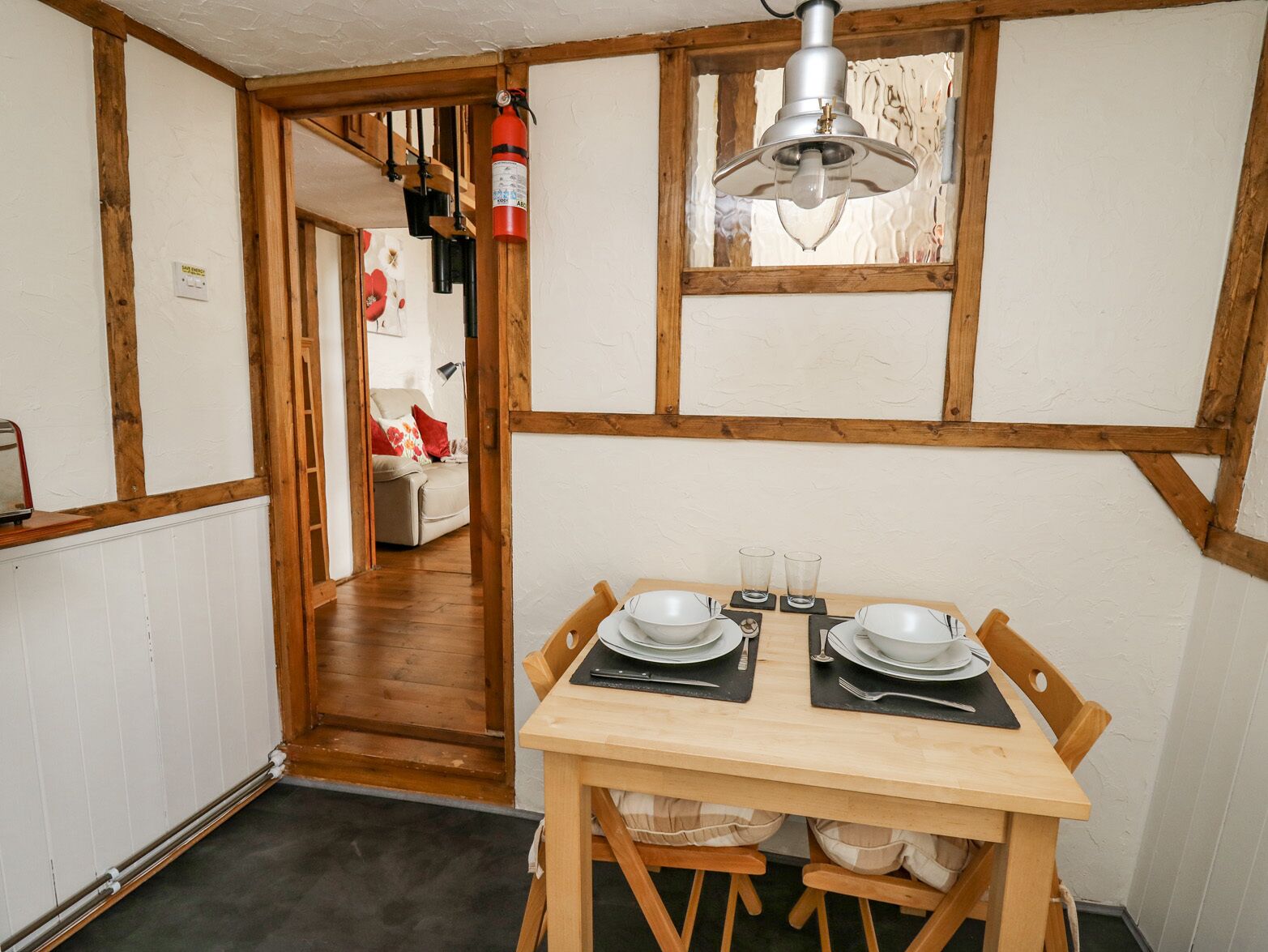 Uma sala de jantar com uma mesa de madeira posta para dois, um extintor de incêndio e um lustre.