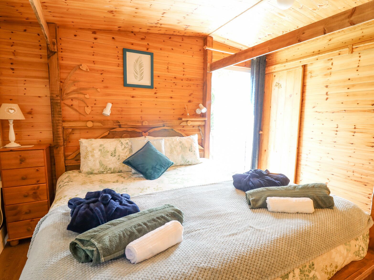 A wooden cabin room with a bed, a nightstand, a framed leaf artwork, and a window.