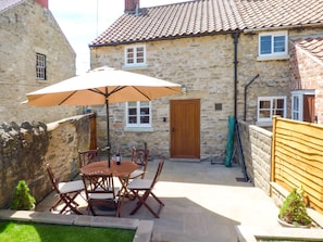 A stone building with a tiled roof, a wooden door, and a small window. There is a table with chairs and an umbrella outside.