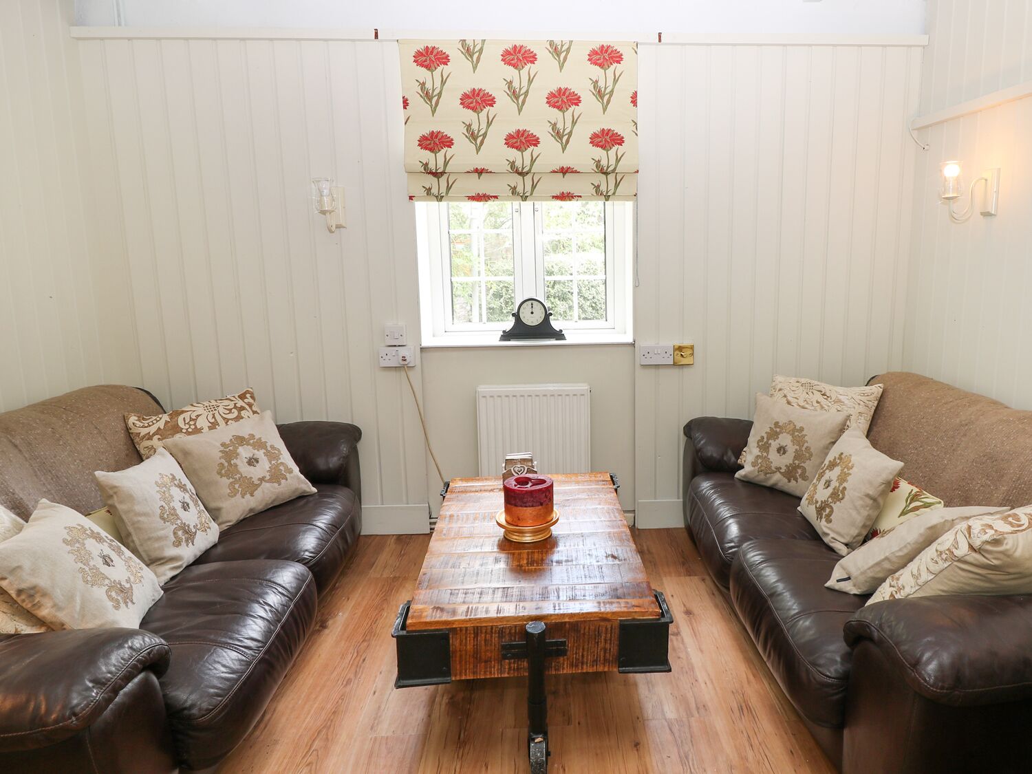 A living room with two leather sofas, a wooden coffee table, a window with a floral valance, and a wall-mounted clock.