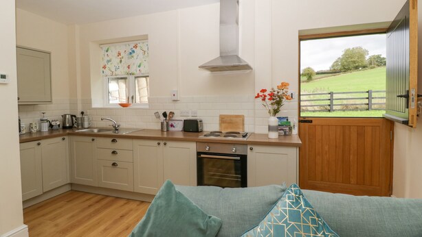 Una cocina con armarios de madera, horno empotrado, ventana con cortina floral y vista a un campo verde a través de una puerta abierta.