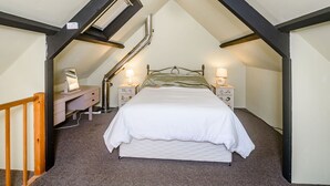 A bedroom with a sloped ceiling, a bed with white bedding, two bedside tables with lamps, and a small desk with a mirror. - Creenagh's Cottage (Minehead)