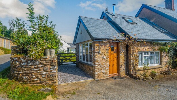 A stone cottage with a wooden door, a gravel driveway, and a stone wall with plants. - Creenagh's Cottage (Minehead)