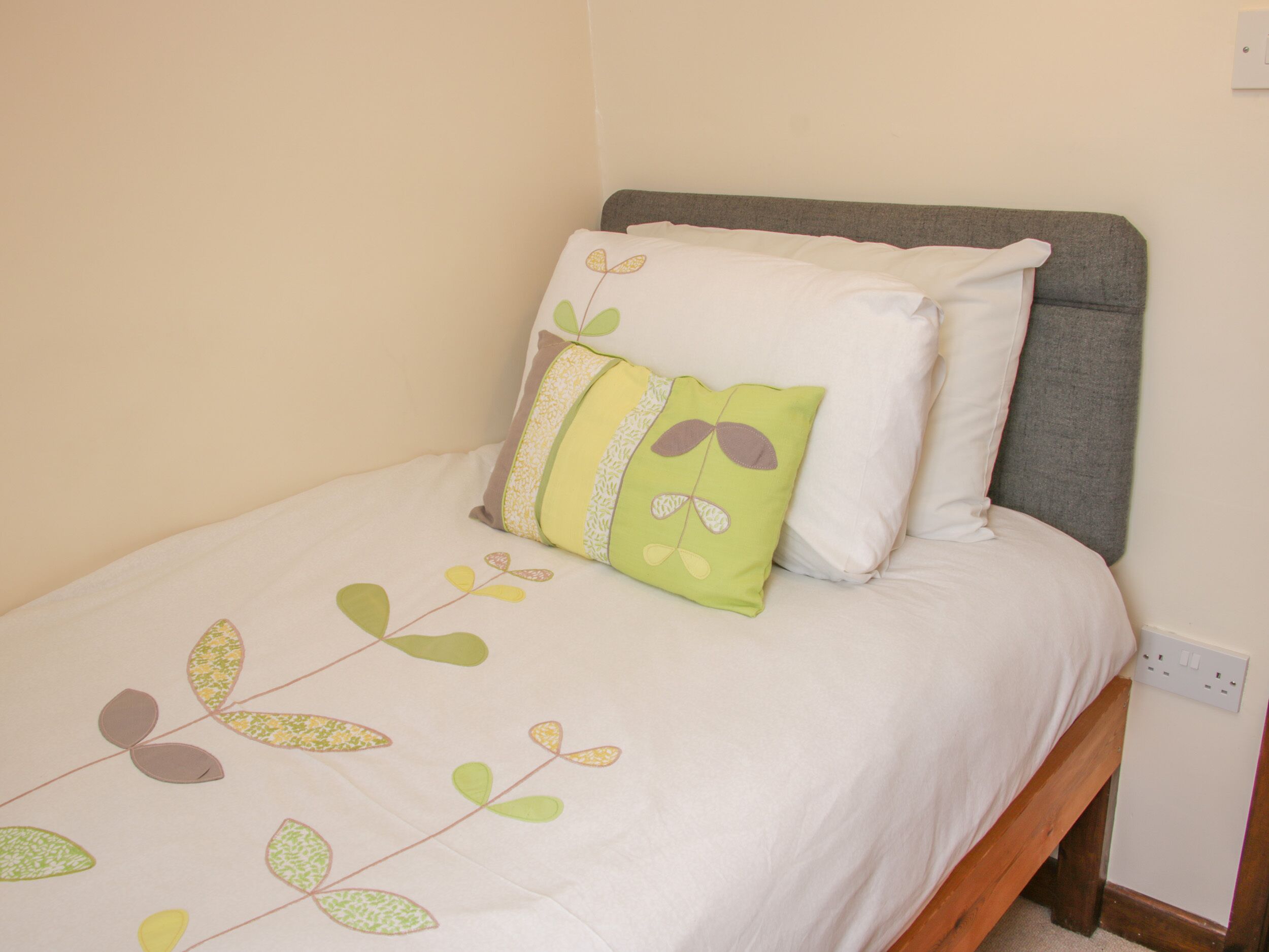 Una cama individual con cabecera gris, cubrecolchón blanco y almohada verde con estampado de hojas.