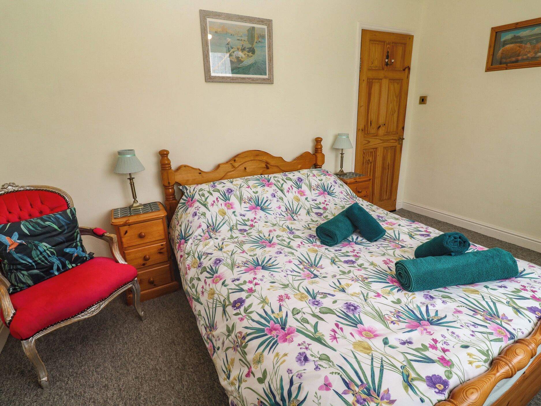 A bedroom with a floral bedspread, a wooden headboard, a red upholstered chair, a wooden nightstand with a lamp, and a wooden wardrobe.