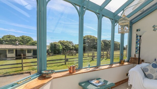 A conservatory with large windows, a wicker table, a bed, and a view of a rural landscape.