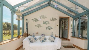 A spacious room with a high ceiling, a white sofa with patterned cushions, a bench, and a view of a green landscape through large windows.