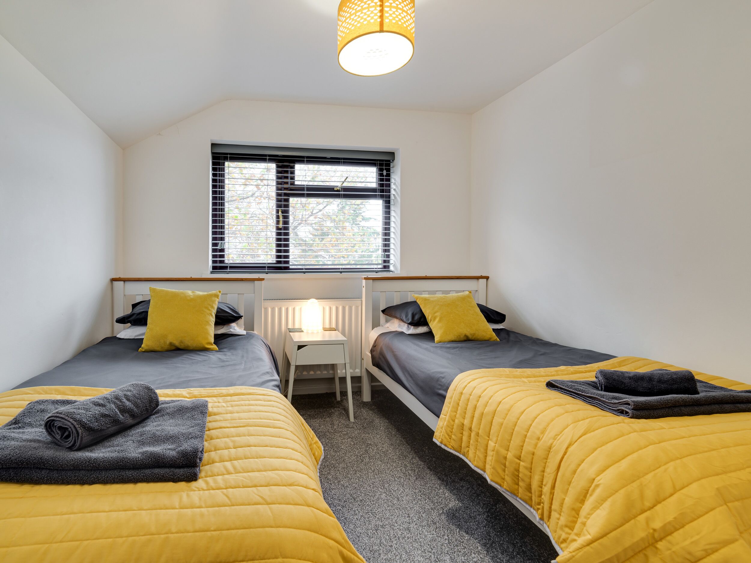 A bedroom with two beds, each with a yellow duvet and a grey blanket, a small desk with a lamp, and a window with blinds.