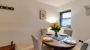 A dining area with a round wooden table set for four, surrounded by grey upholstered chairs. A vase with yellow flowers is on the table, and there is a framed picture on the wall above it. - 2 Ysgoldy (Bangor)
