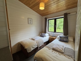 Un dormitorio con dos camas, techo de madera, ventana con cortinas y una mesita de noche con lámpara.