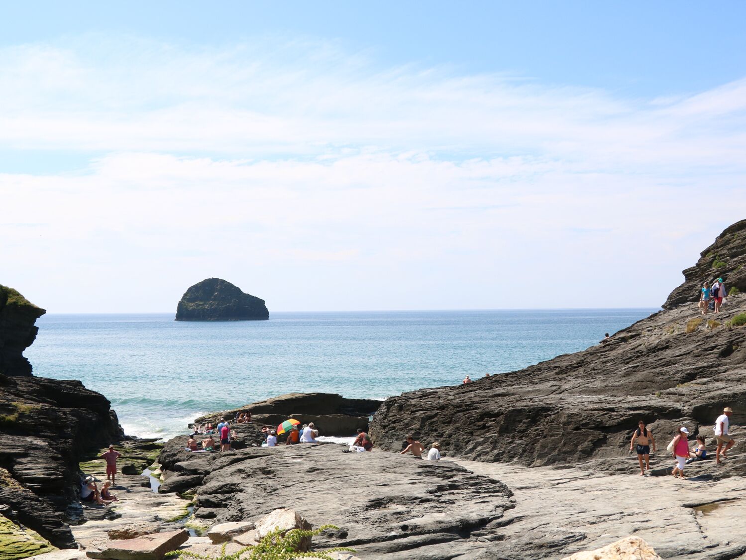 A rocky beach with a large rock formation in the distance, surrounded by clear blue water and a few people enjoying the area.