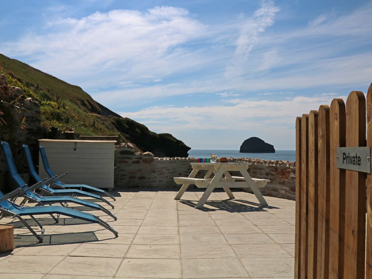 Un patio privado con sillones azules, una mesa de picnic y un muro de piedra con vistas al mar.