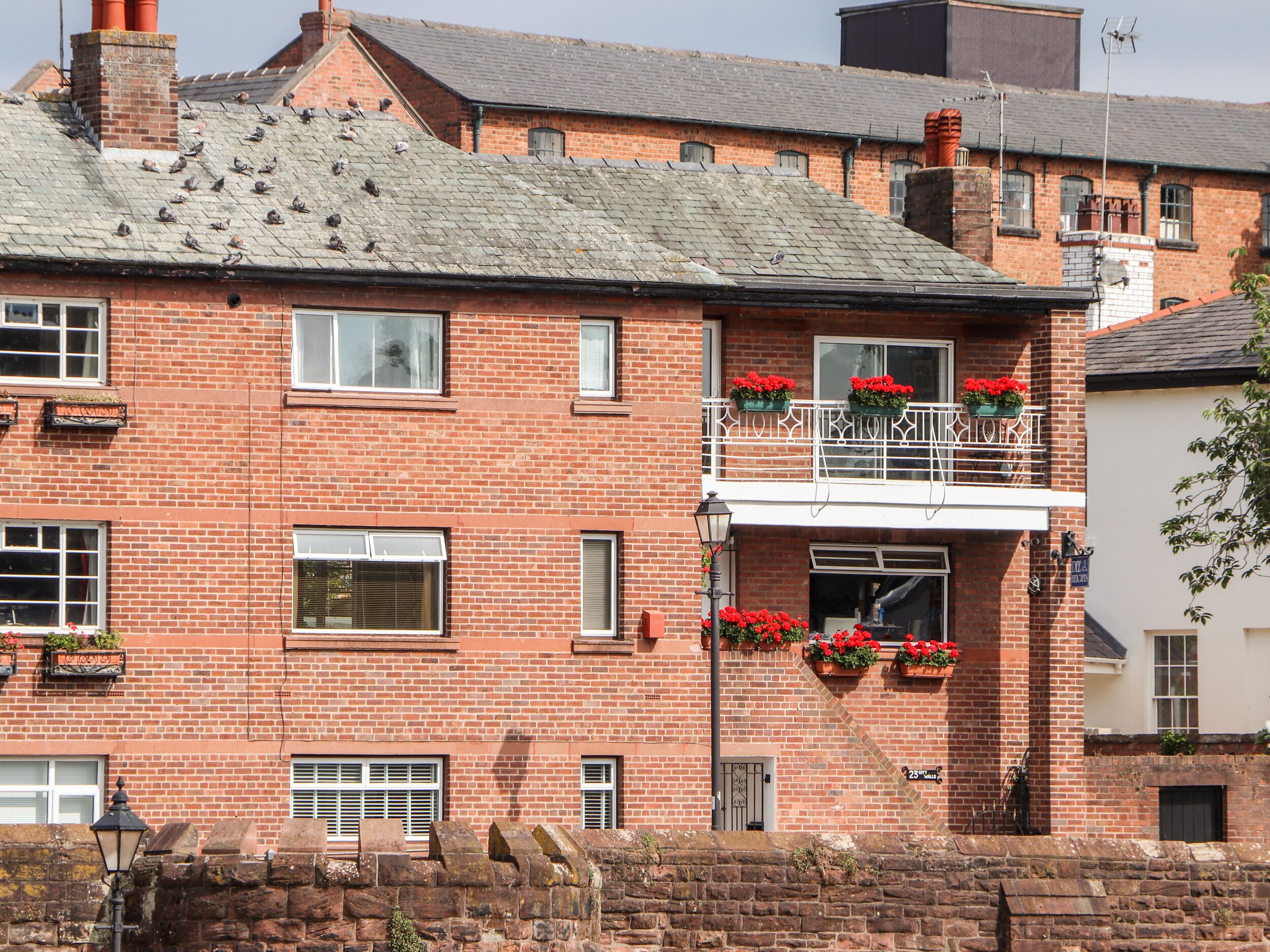 A red brick building with multiple windows, a balcony with flower boxes, and a stone wall in the foreground.
