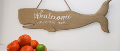 Placa de madeira em formato de peixe com o texto 'Whalecome to our beach house' pendurada numa parede.