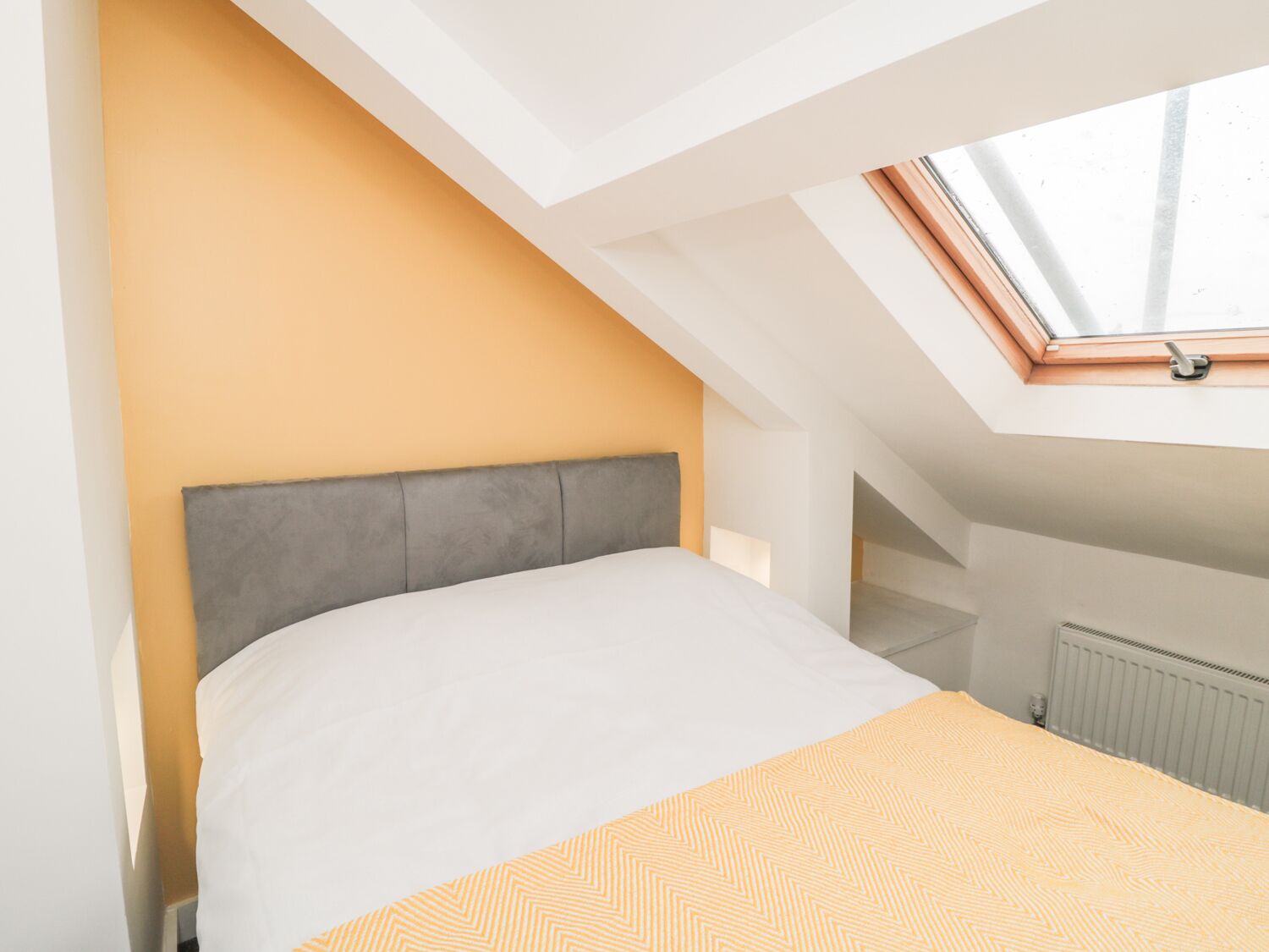 A bedroom with a sloped ceiling, a skylight, a bed with a grey headboard, and a radiator.