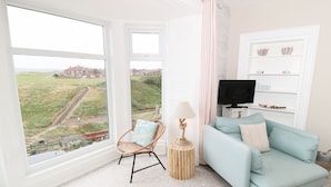 A room with a large window offering a view of a residential area, a light blue sofa, a wicker chair, a small round table with a lamp, and a television on a white shelf. - Larksbay View (Redcar)