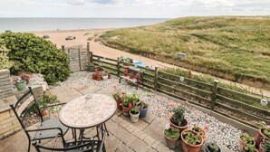 A patio with a mosaic table, chairs, and potted plants overlooking a beach and dunes. - Larksbay View (Redcar)