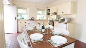 Une cuisine bien éclairée, avec des meubles blancs, une table à manger dressée pour quatre personnes et une horloge au mur.