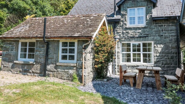 Une maison en pierre avec un toit en tuiles, un petit jardin avec du gravier, et une table et des chaises en bois.