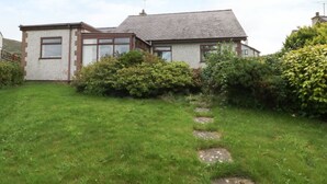 Une maison à deux étages, dotée d’un toit en pente, d’une cheminée bien visible et d’un petit jardin avec un sentier en pierre menant à l’entrée.