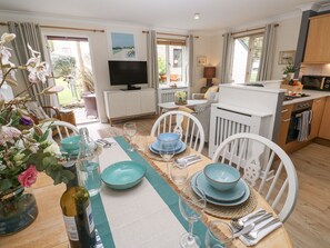 Un coin repas avec une table dressée pour quatre personnes, un téléviseur en arrière-plan et une cuisine visible sur la droite.