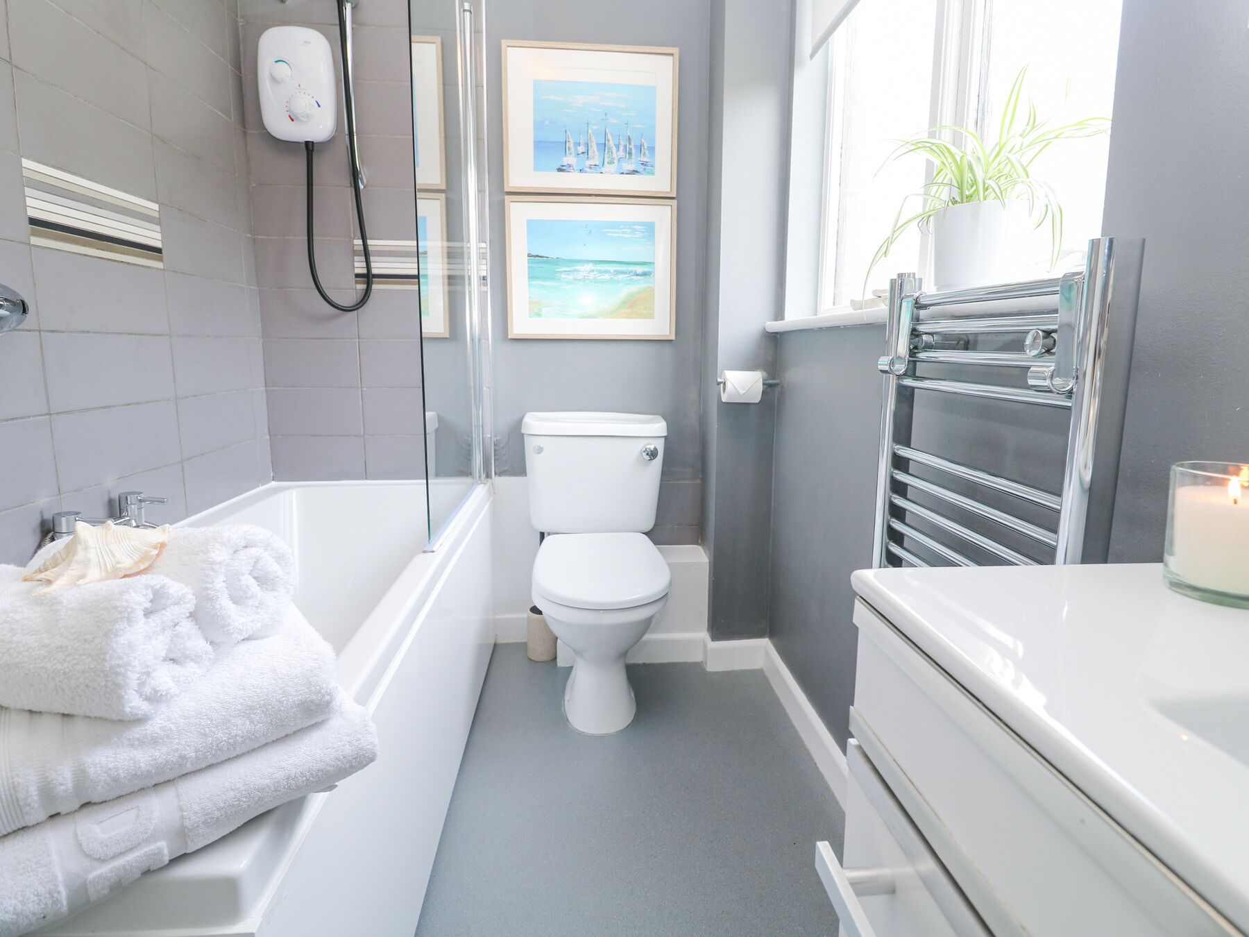 A modern bathroom with a bathtub, toilet, and sink. There are framed pictures on the wall, a towel rack, and a plant by the window.