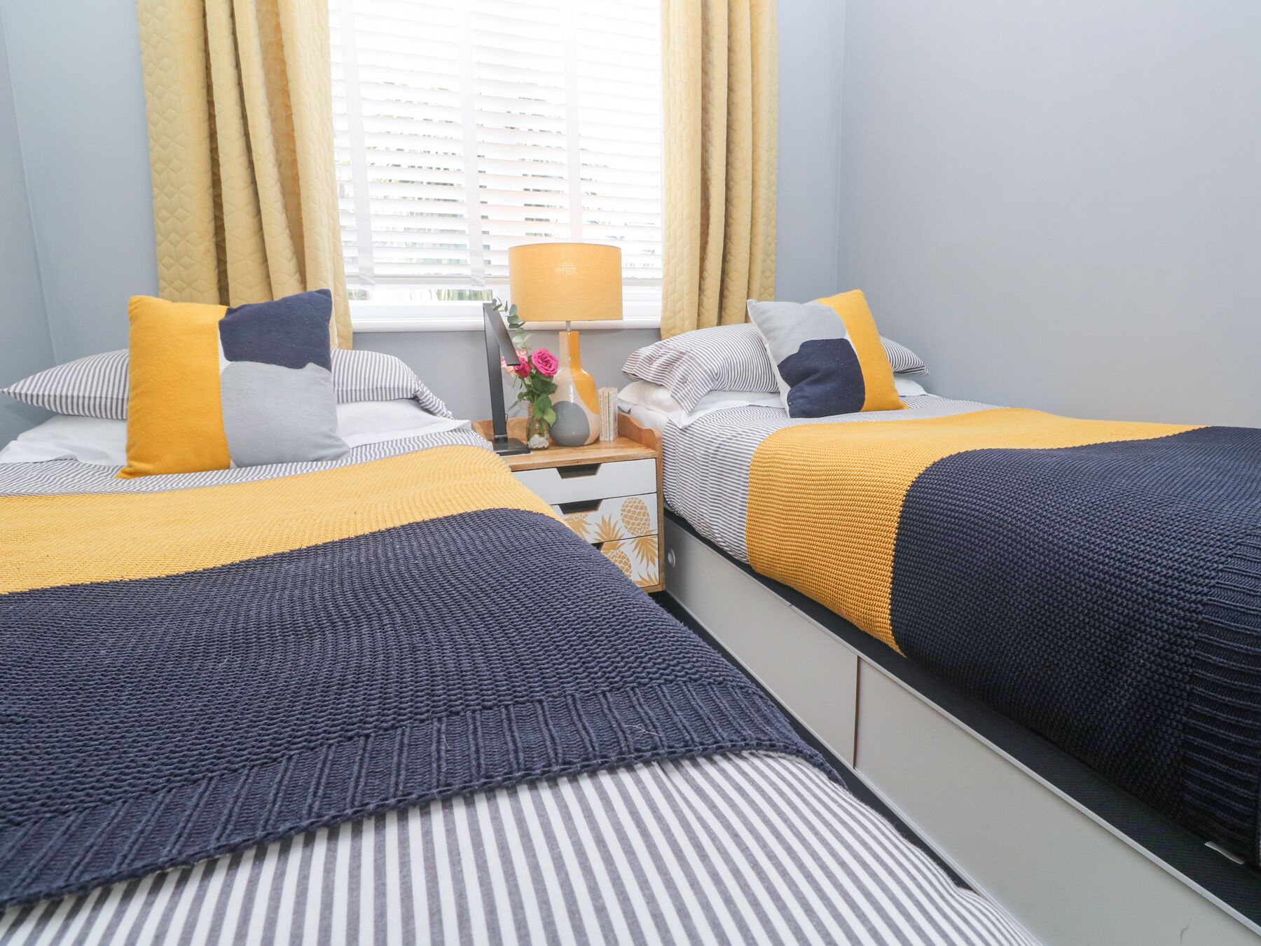 A bedroom with two beds, each with striped bedding and a yellow accent pillow. There is a nightstand with a lamp and a vase with flowers.