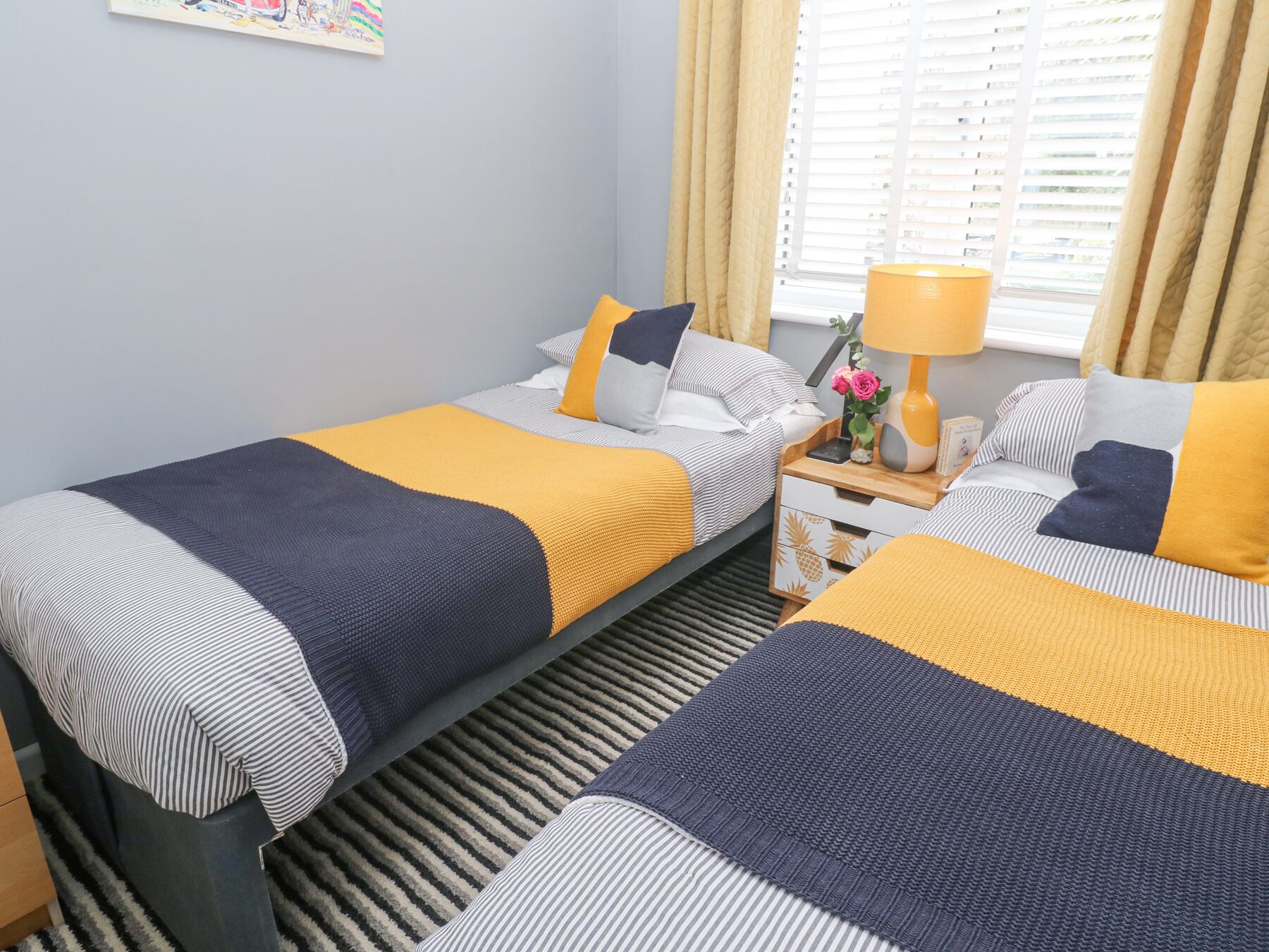 A hotel room with two beds, each with striped bedding and a yellow accent. There is a nightstand with a lamp and a vase of flowers.