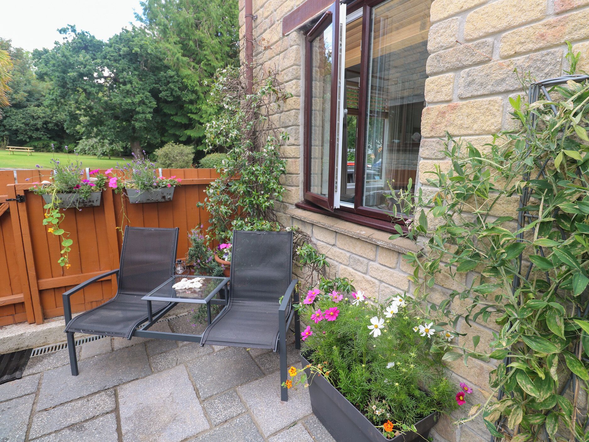 A patio with two black mesh chairs, a glass table, and a stone floor. There are flower planters with various plants and flowers.