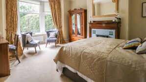A bedroom with a large bed, a wooden wardrobe, a chair, a fireplace, and a window with curtains. - The Garden Flat (Harrogate)