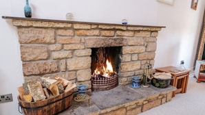 Ein Kamin aus Stein mit Feuer, ein Weidenkorb für Holz und ein kleiner Tisch mit Büchern und einer Vase.