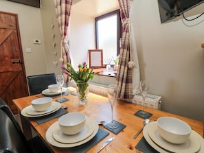 Una mesa de comedor dispuesta con platos, tazones y copas de vino blancos, un florero con flores y cortinas a cuadros.