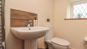 A bathroom with a white sink, a wooden panel above the sink, a toilet, a small trash bin, and a window with a plant.