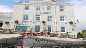 Un bâtiment blanc avec une enseigne indiquant «  Pier House » et « Stanstell Brewery » sur la façade. Des jardinières remplies de fleurs sont posées sur les rebords des fenêtres, et un bateau nommé « Rashleigh » est amarré devant.