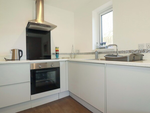 A modern kitchen with white cabinets, a stainless steel oven, a black microwave, a kettle, a window, and a tiled backsplash. - Little Pippin (St Austell)