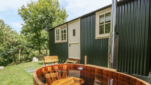 A wooden hot tub with a glass enclosure, a wooden deck, and a green building with a door and windows. - Orchard (Wadebridge)