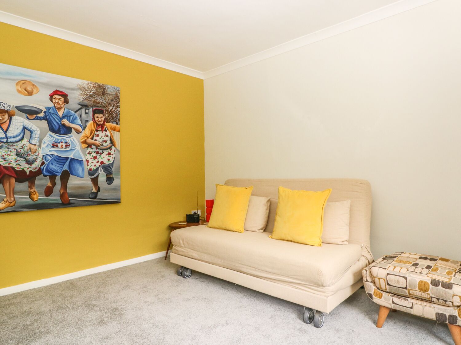 A room with a yellow wall, a beige sofa with yellow cushions, a small ottoman, and a painting of people in vintage clothing.