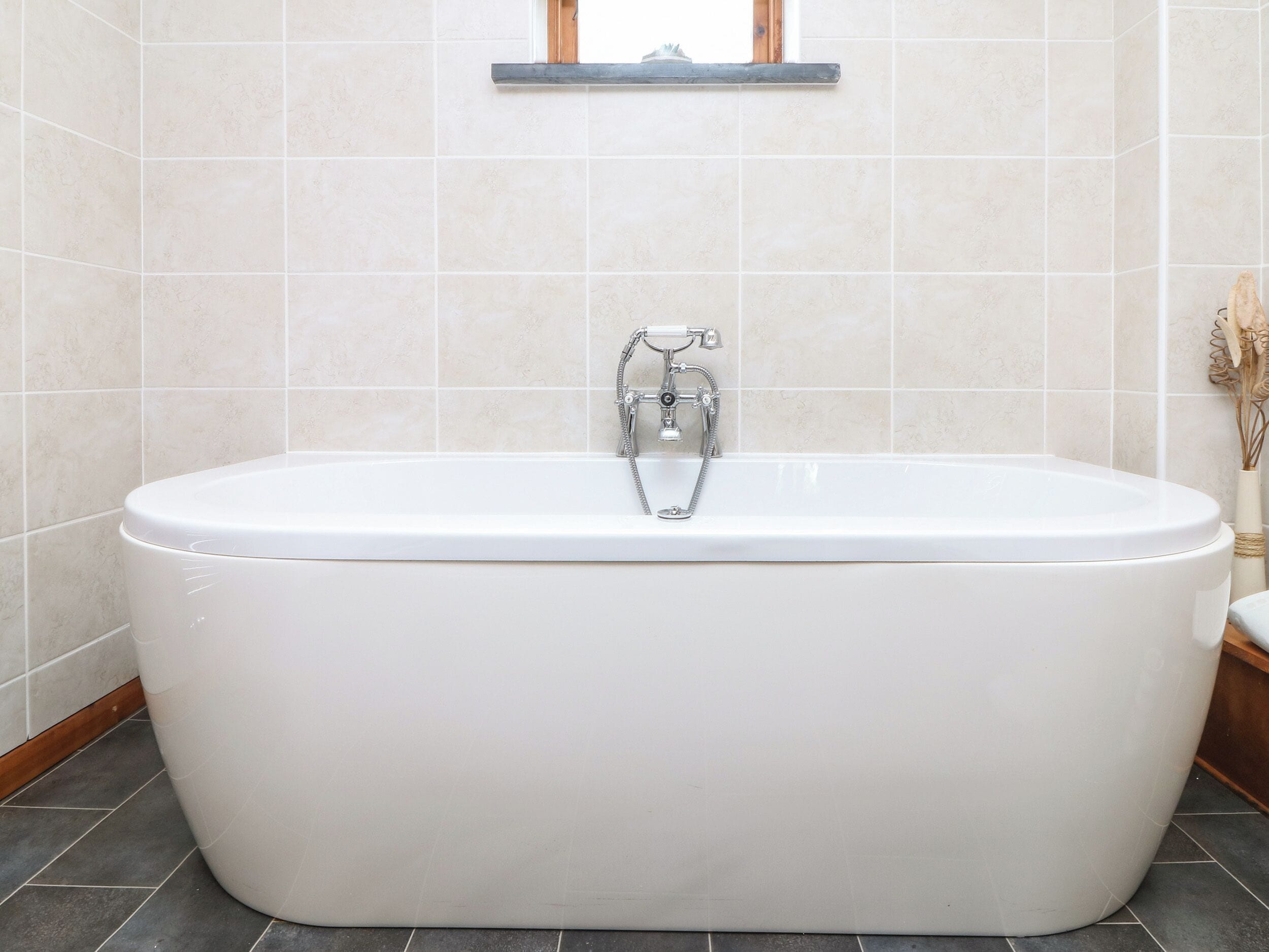 A modern bathroom with a freestanding bathtub, a window above the tub, and a vase with dried flowers on the right.
