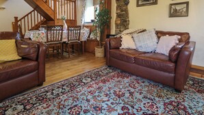 A cozy living room with a brown leather sofa, a dining table with floral tablecloths, and a staircase with wooden beams. - The Croft (Holsworthy)